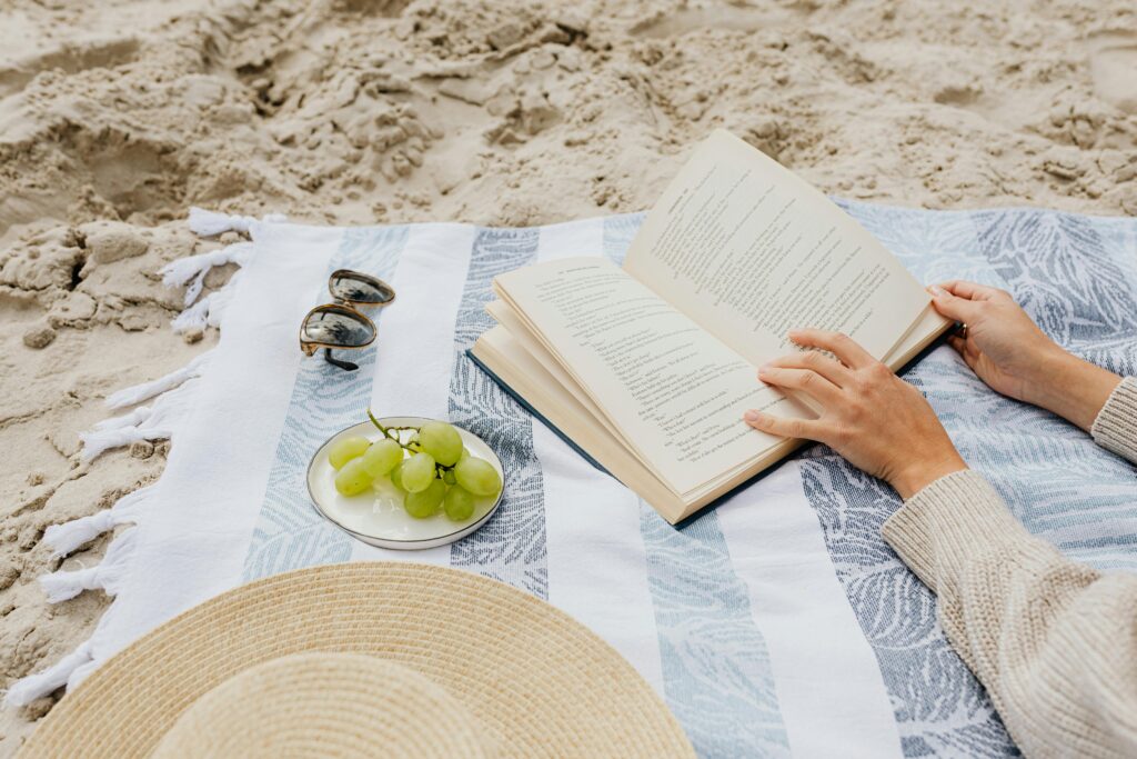 “5 Must-Read Summer Beach Books for a Perfect Vacation (2026)” Beachside picnic scene with book, green grapes, and sun hat on a blanket.